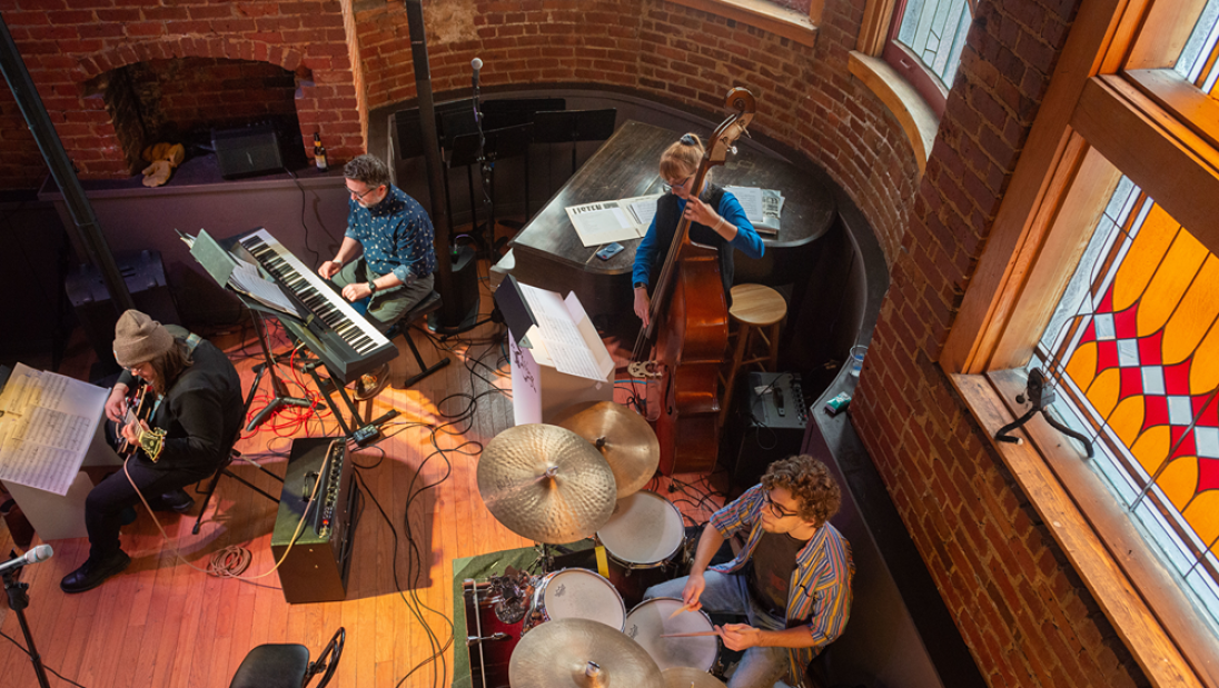 A jazz band performing in a cozy, brick-walled venue with stained glass windows, featuring musicians on keyboard, guitar, double bass, and drums.