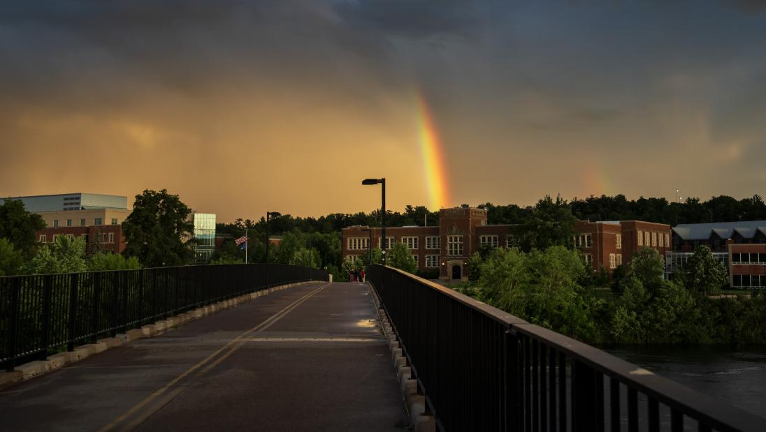 A rainbow emerges from the clouds and appears to end on Schofield Hall as seen from the footbridge.