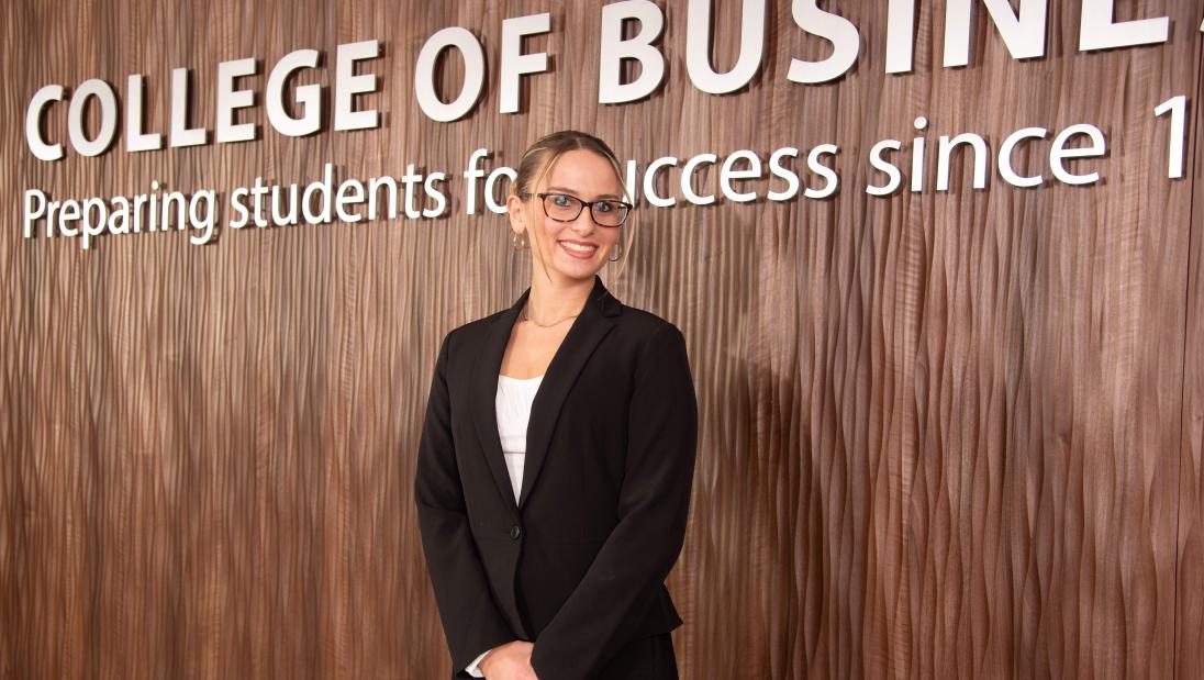 Departmental Honors student in Marketing, Kaitlyn Riehle, standing in front of the College of Business lobby sign. 