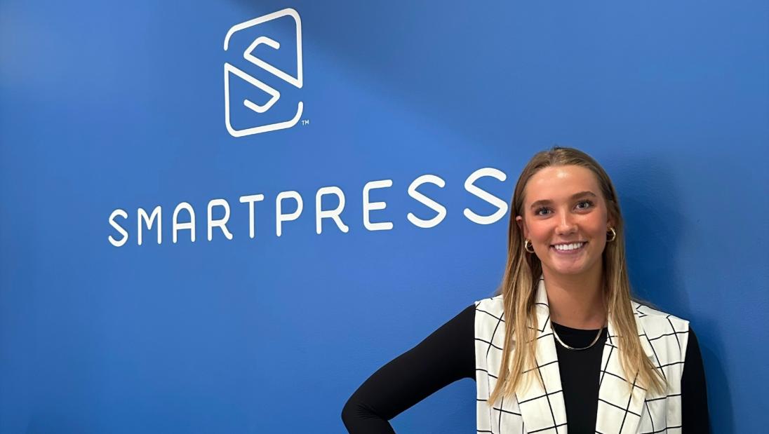 Marketing student, Lauren Schubert, standing next to company logo at her internship with Smartpress.