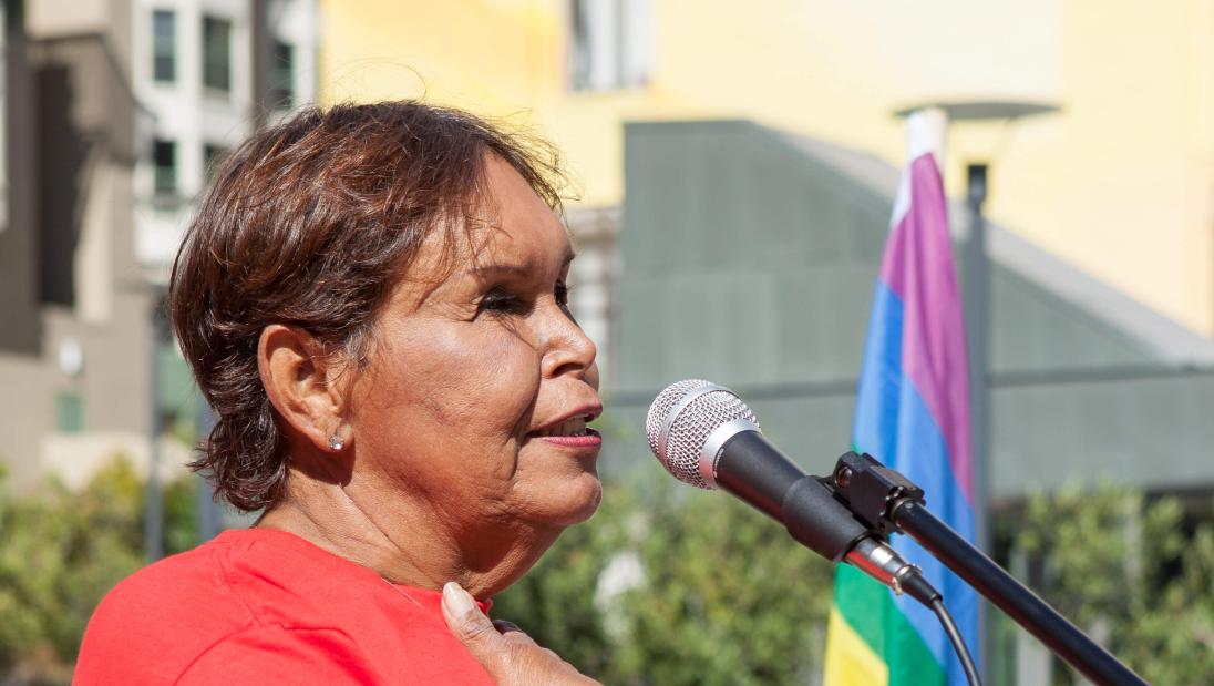 A woman wearing a red shirt, with her hand on her heart, stands aside a rainbow flag.