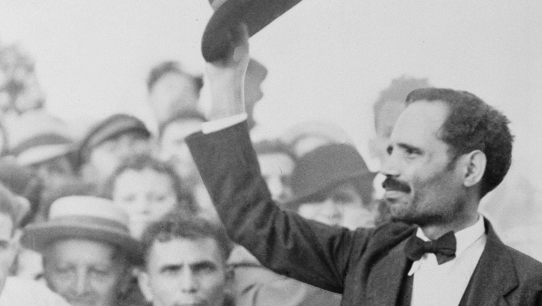 Black and white photograph of a man raising his hat to a crowd of onlookers.