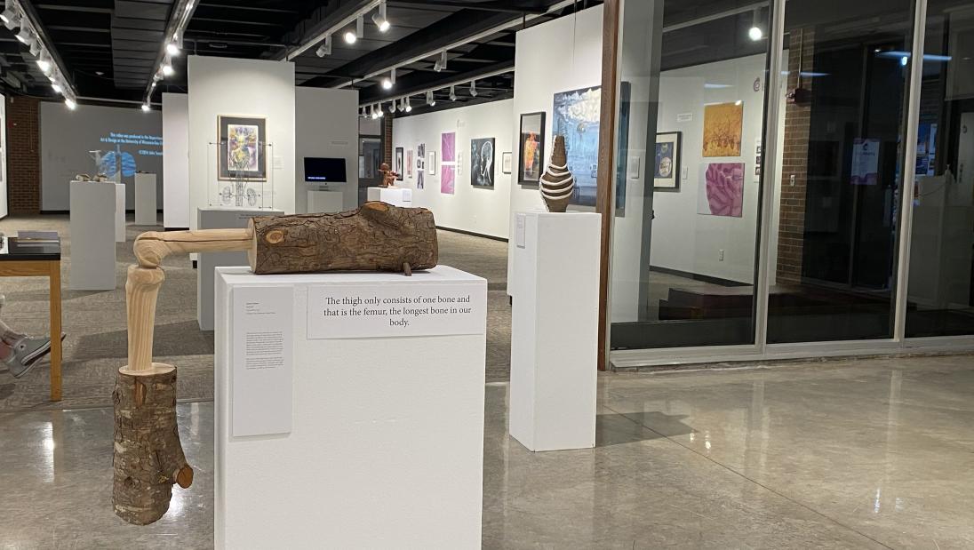 Installation view of Body Parts exhibition. In the foreground a sculpture sits on a pedestal. A bone form emerges from a log, hanging at a ninety degree angle, another bone emerges from a log hanging down the side of the pedestal.