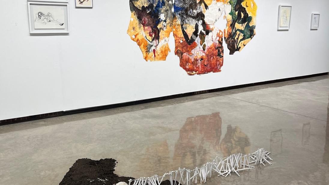 Mixed media sculpture primarily using soil lays on the ground while a chain of suringes stretches out across the floor. A large drawing with orange and yellow hues hangs on the wall behind the sculpture.