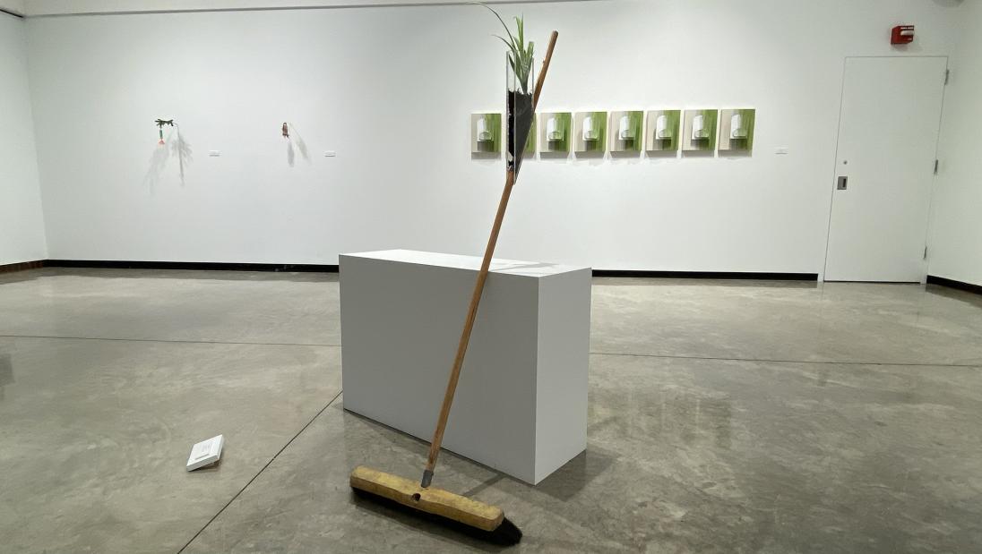 A janitors broom leans against a white pedestal. An acrylic container is attached to the broom handle and a spider plant grows from the container. 