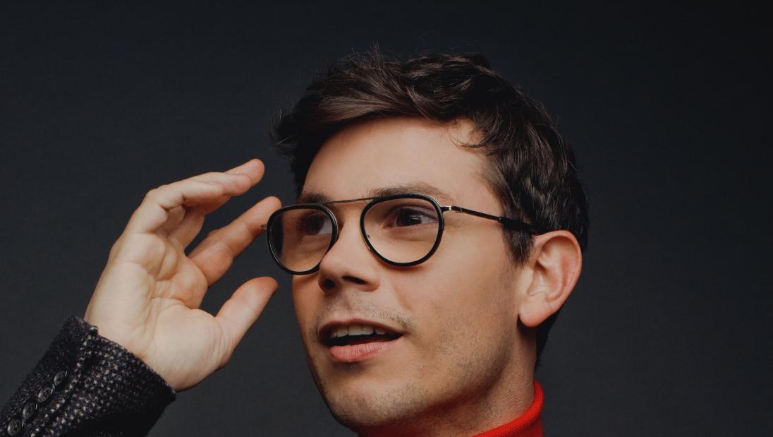 A man, smiles in profile, hand on his glasses.