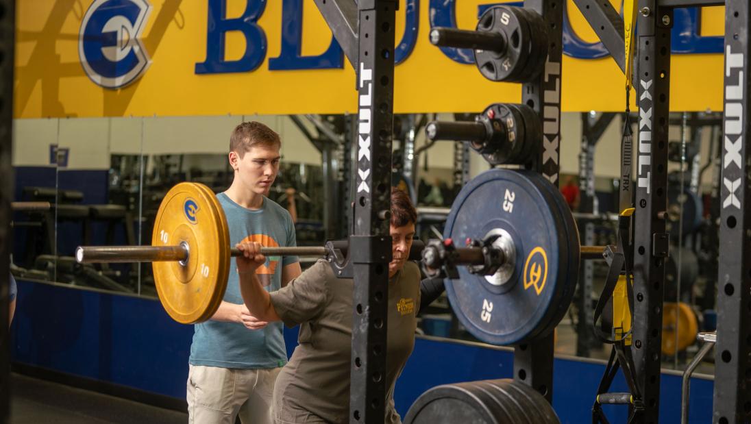 student spotting a client doing a squat in the weight room