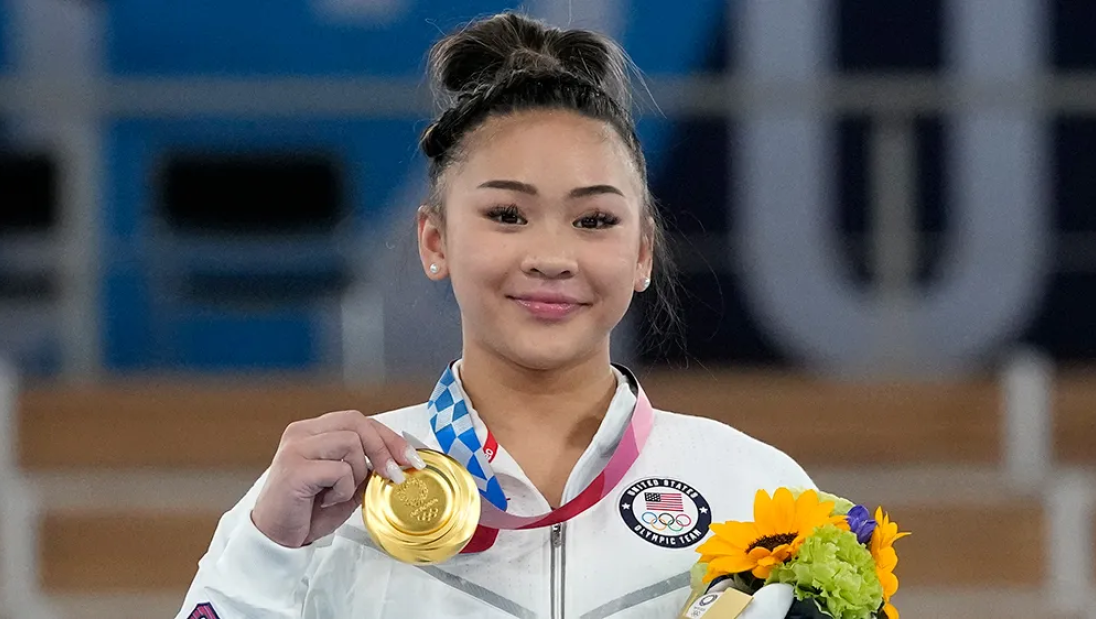A woman smiles for the camera, holding up an Olympic gold medal.