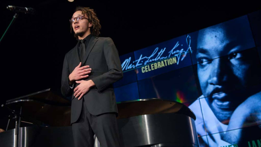 A student, mid-musical performance, stands on a stage in front of a piano with a graphic of Martin Luther King Jr. behind them