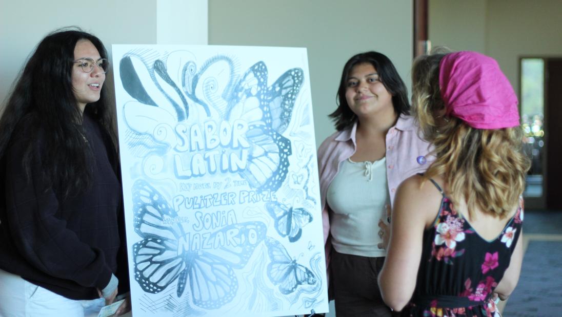 Three students chat while standing by a 'Sabor Latin(Eau)' posterboard 
