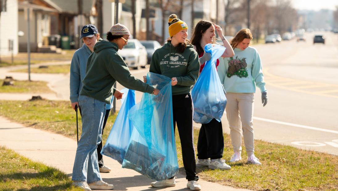 A group of five students holds bags near the side of the road as they pick up trash.