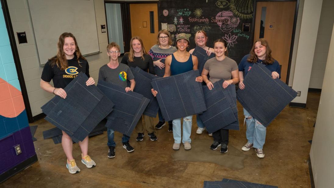 A group of students holds carpet tiles which they pried up from the floor to be recycled. 