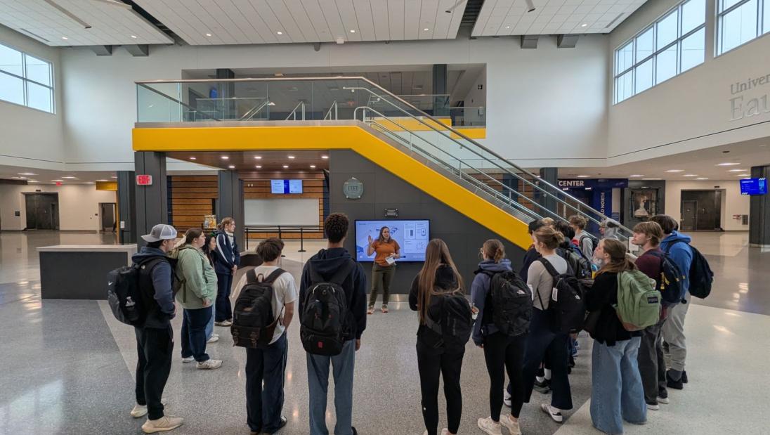 A class of students circles up around a tour lead, who stands in front of the energy dashboard in the lobby of the Sonnentag.