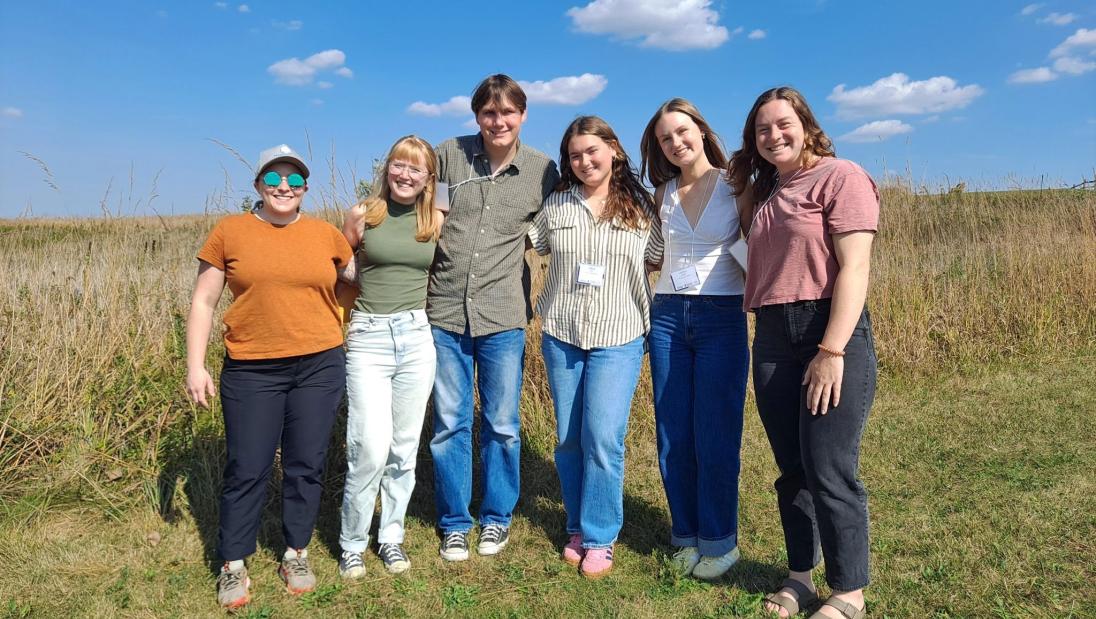 A group of five students stands with their advisor in a prairie, smiling in the sun.