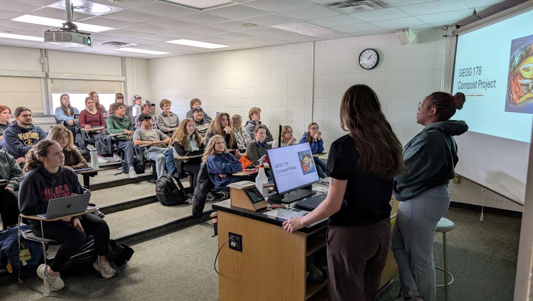 Two students present to a classroom next to a projected slide about compost.