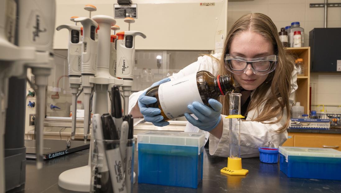 A student pours liquid into a glass beaker while wearing a lab coat, gloves, and safety goggles.