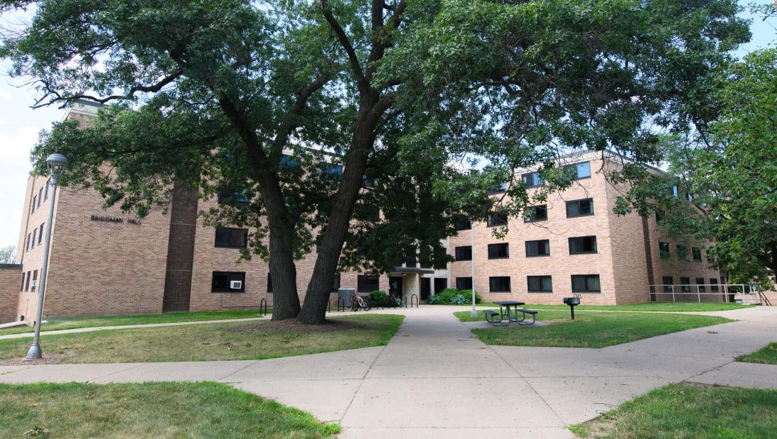 Exterior view of Bridgman Hall, a brick building with lots of windows and a big tree and picnic table in front.