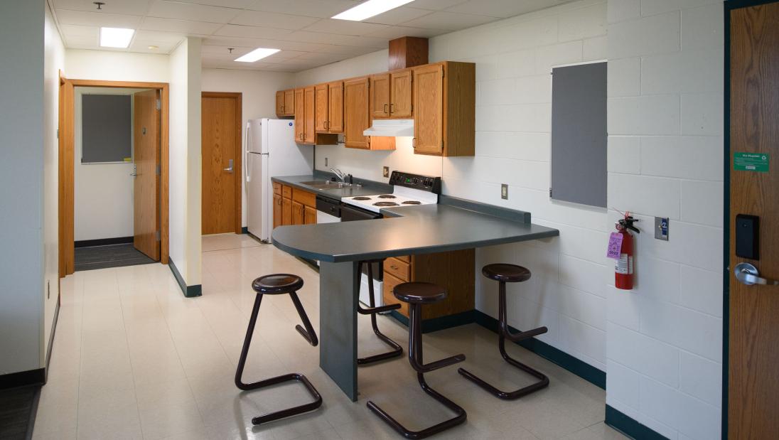 Kitchen area in a Chancellors Hall appartment with fridge, dishwasher, sink, oven, and counter with stools.