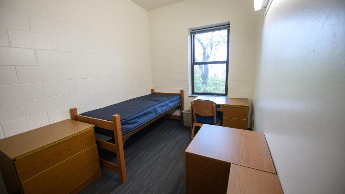 Single bedroom in Chancellors Hall apartment with bed, window, desk, chair, bookshelves, and dresser.