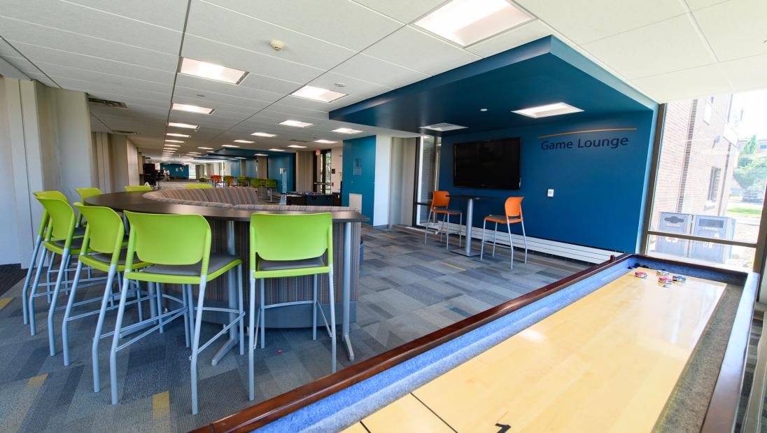Game room in Towers Hall with colorful chairs, shuffleboard, and large TV.