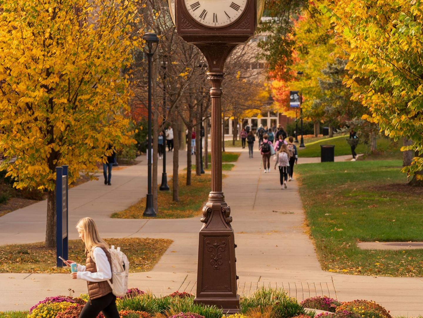 Student walks across campus in autumn in front of clock.