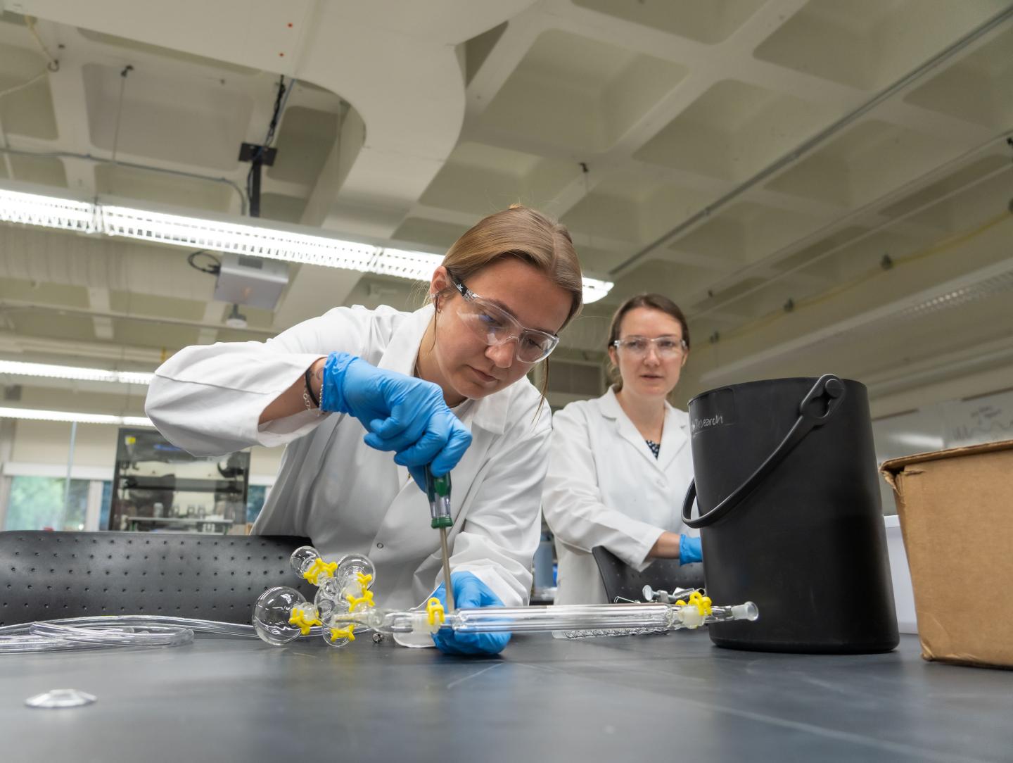 Two students wearing white lab coats work together connecting a piece of tubing in a lab setting 