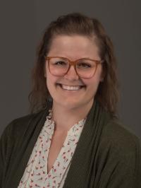 Person with glasses wearing a green cardigan and floral blouse against a gray background.