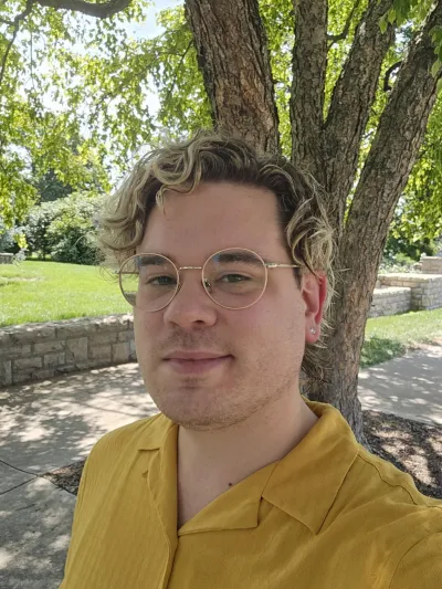 An image of Professor Chris Outzen, a white cisgender man in his mid-30s with blonde hair, in a yellow shirt under a tree.