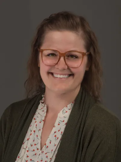 Person with glasses wearing a green cardigan and floral blouse against a gray background.