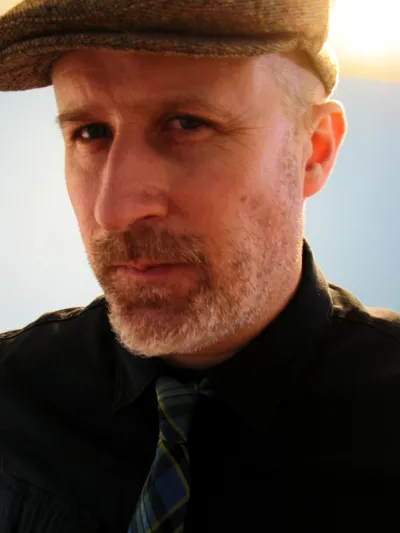Image of Ned Gannon in a Tweed Hat and Plaid Tie.