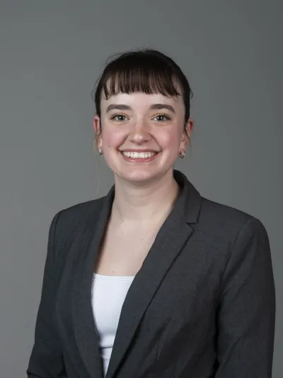 Image of a grey background, young white woman with brown hair, a white shirt, and grey blazer smiling at the camera.
