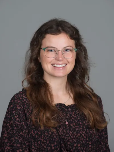 A smiling young woman with long, wavy brown hair and green-framed glasses, wearing a black floral-patterned top against a gray background.