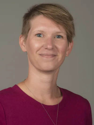 Katie Larson appears smiling. She has short blonde hair and is wearing a maroon dress. 