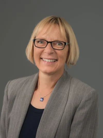 Headshot of woman with blue glasses and gray blazer.