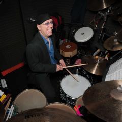Man wearing glasses, a black hat and a blue shirt holds a pair of drumsticks while sitting at a drum set.