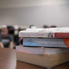 A stack of leadership books on a table with students sitting and talking in the background.