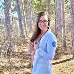 female nursing student in white coat, outside in wooded backdrop 