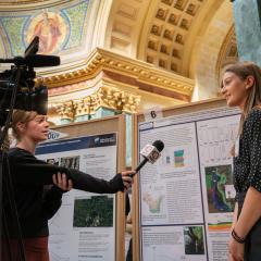 A student researcher being interviewed by a news crew in front of her research poster during the annual research in the Rotunda event