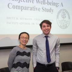 A student and a professor pose and smile in front of a PowerPoint screen while presenting their research on well-being.