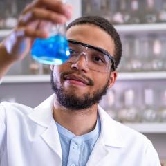 A student examines a beaker full of blue liquid while wearing a lab coat and safety glasses.