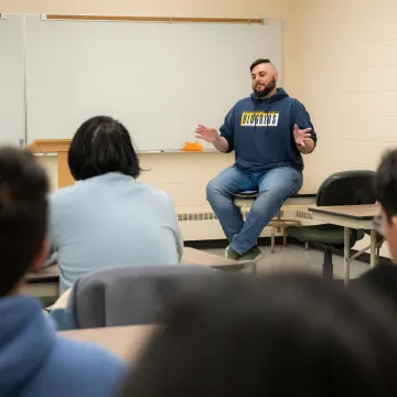 instructor seated on top of a table in a classroom facing students in desks 