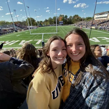 Two students pose in the stands with the football field behind them.