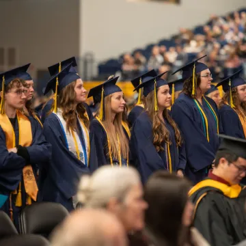 Students wearing graduation gowns stand and watch the ceremony