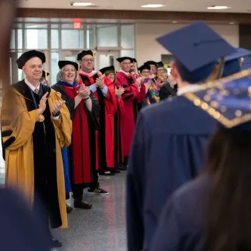 Graduates wearing graduation caps walk in a line to cheers from faculty