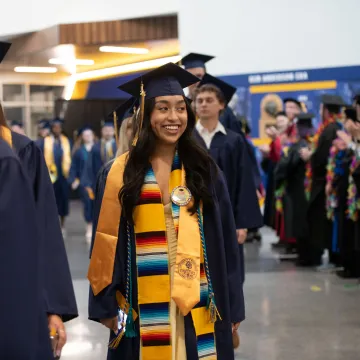 Students wearing graduation caps and gowns smile as they walk in to the ceremony