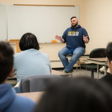 instructor seated on top of a table in a classroom facing students in desks 