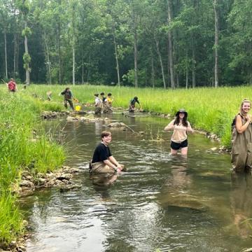 Students wade in a creek and smile while collecting water samples