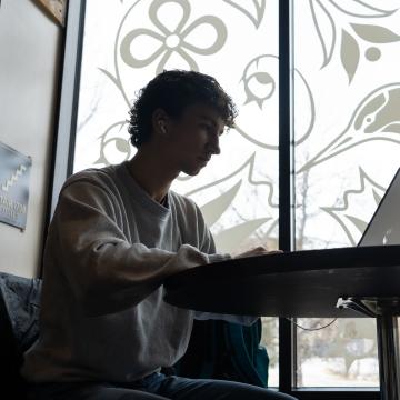 A student studies on his laptop at a table by a window.