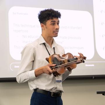 A student holds a small viola while sharing his research findings during CERCA. A display of music notes is in the background.