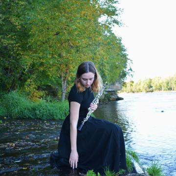 Carrie Schwartz in a black dress balances over a river, oboe in hand 